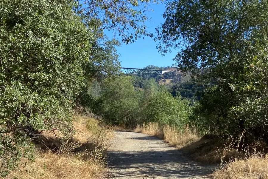 Foresthill Bridge visible beyond a shaded dirt trail above the American River canyon
