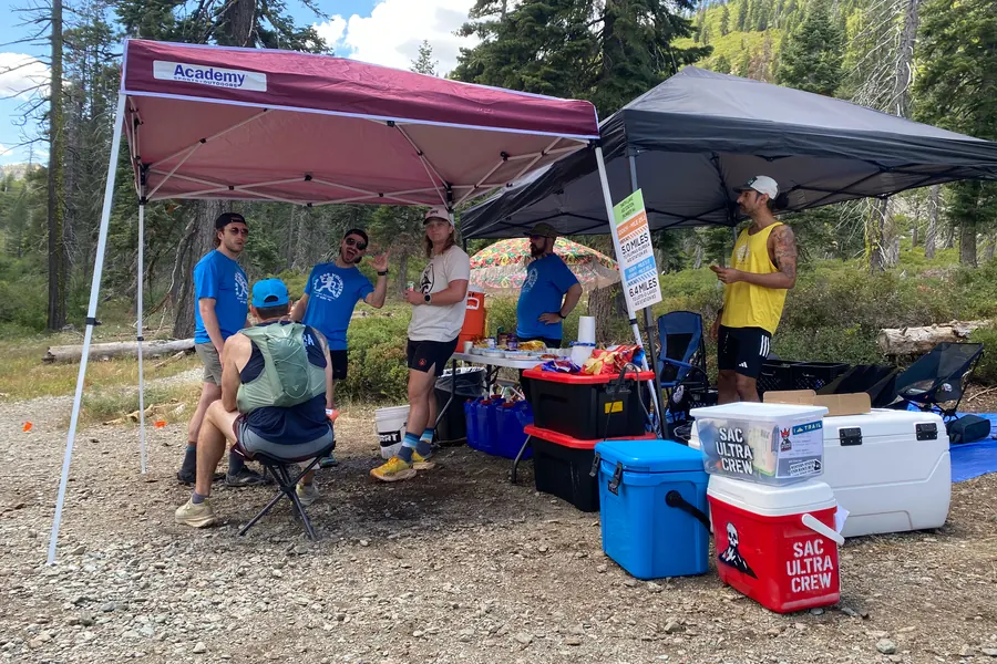 Trail-race aid station set beneath trees with food, water, and Sacramento Ultra Crew coolers